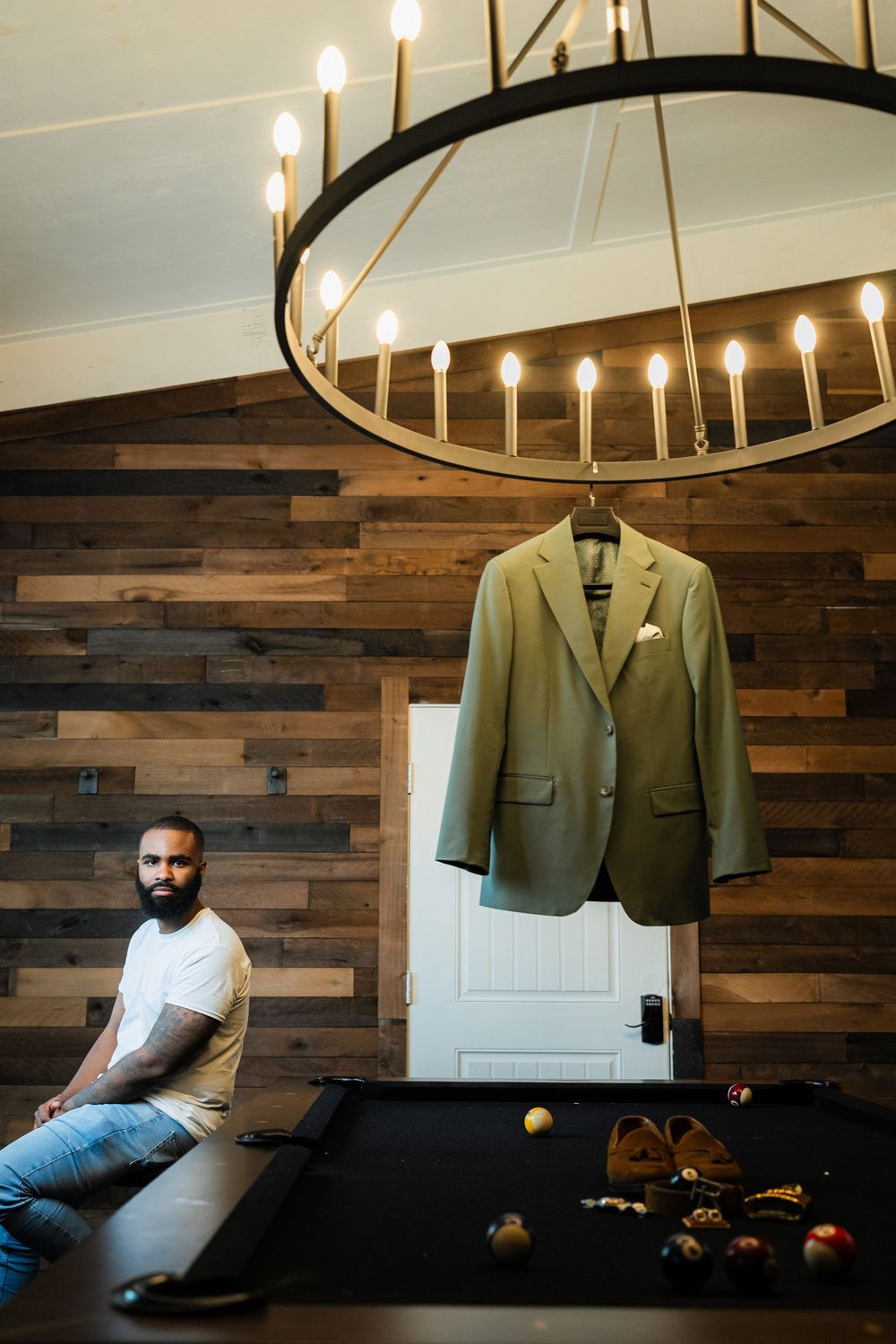 A suit jacket hangs under a modern circular chandelier against a rustic wooden wall backdrop with pool table below.
