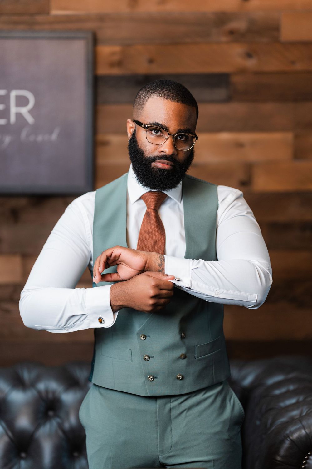 A well-dressed person in a mint vest and coral tie adjusts their sleeve against a wooden wall backdrop.