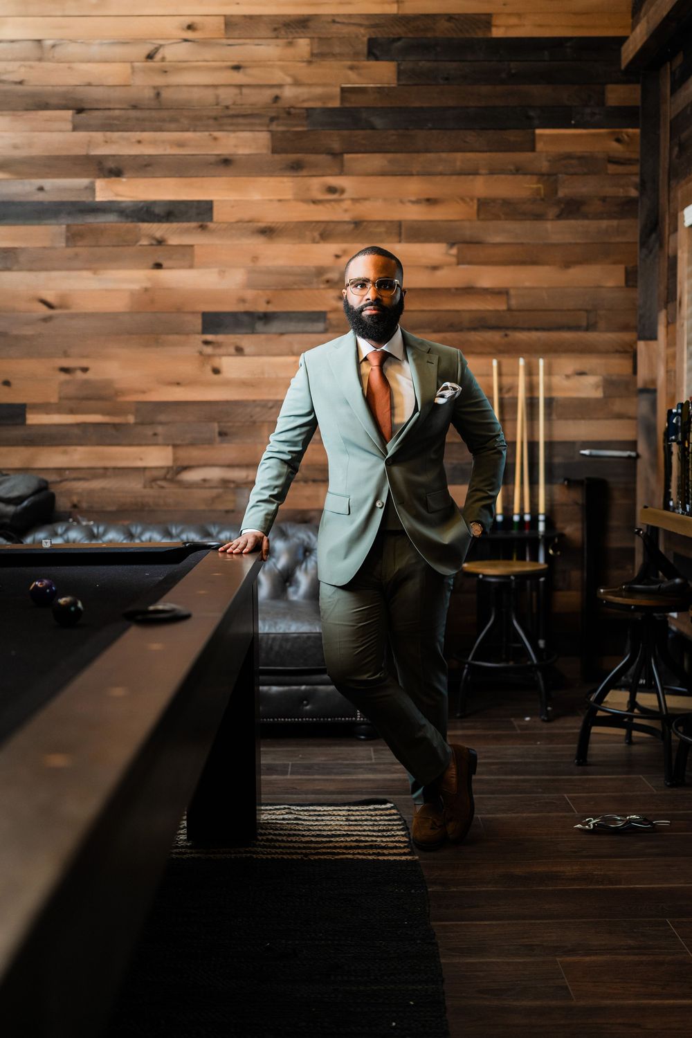Modern sage green suit styled with copper tie in rustic wood-paneled room with pool table.