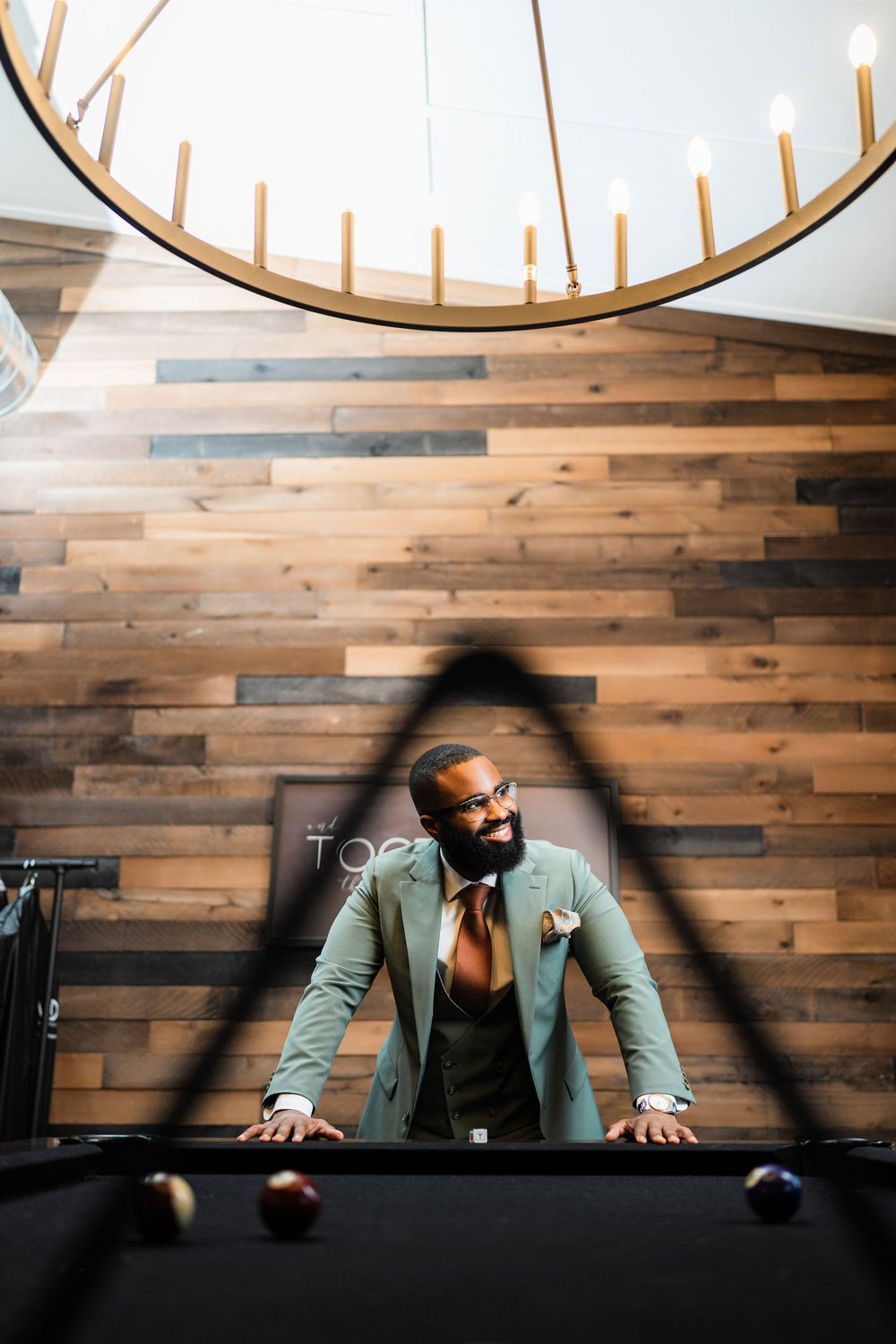 A person in mint green suit leans over pool table under dramatic circular chandelier lighting.