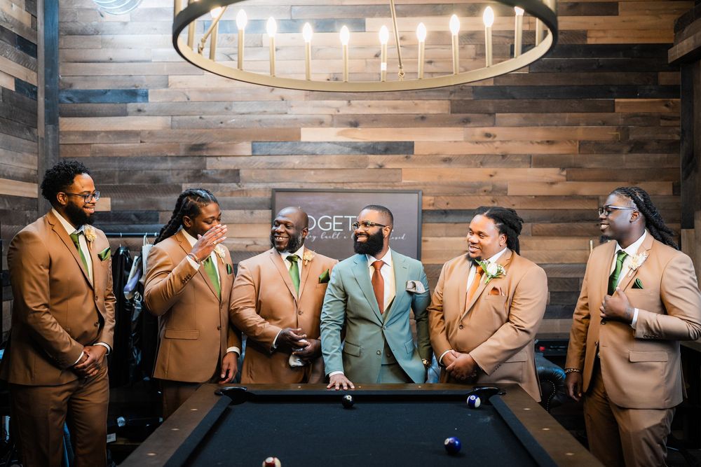 Group gathered around pool table wearing coordinated suits in earth tones with wood paneled wall behind them.