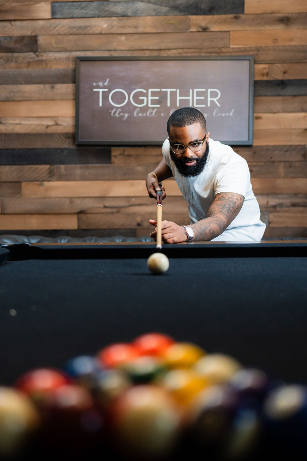 Playing pool in front of rustic wooden wall with 'Together' sign and racked colorful billiard balls in foreground.