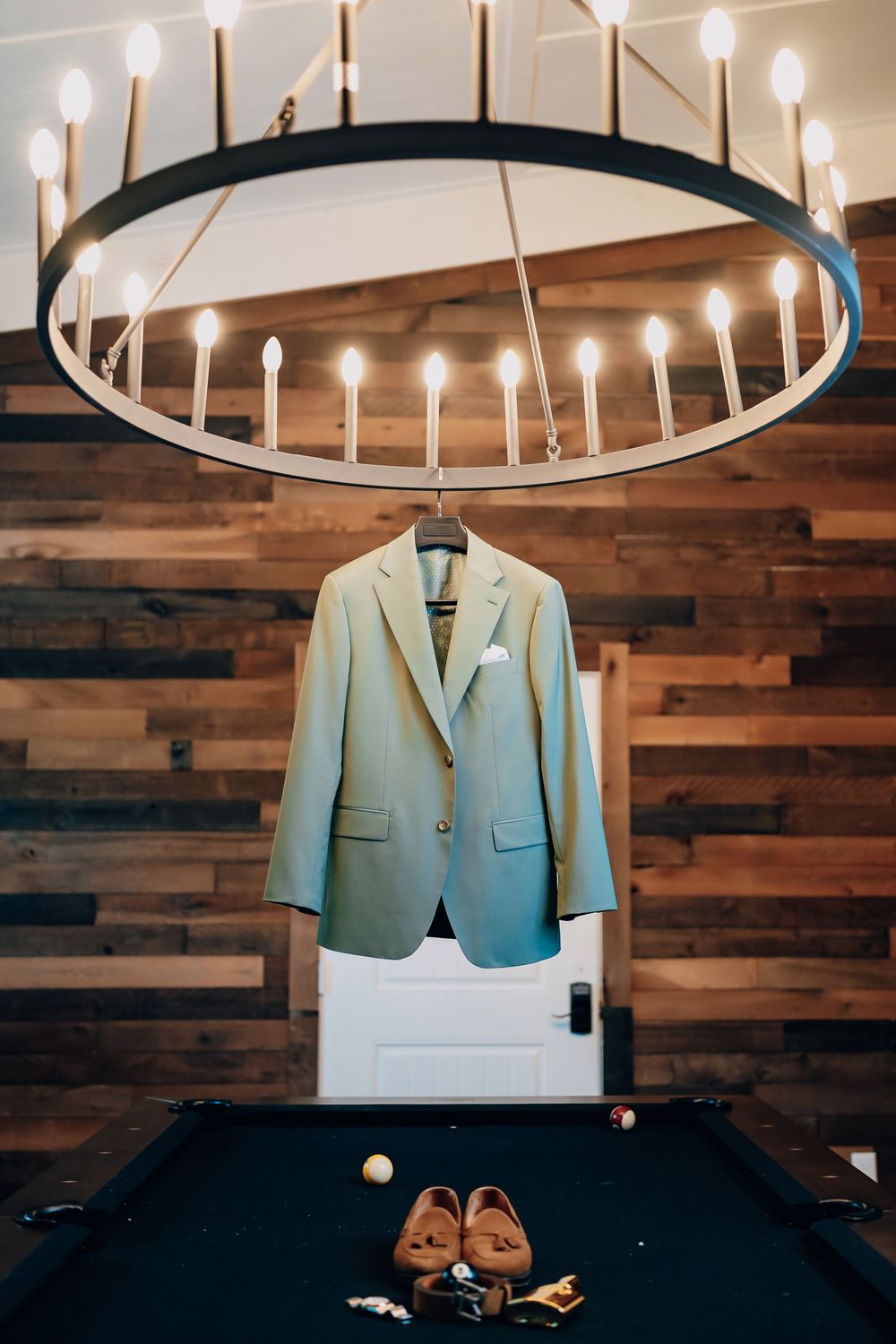 A mint green suit jacket hangs under a modern circular chandelier above a pool table with brown leather shoes.