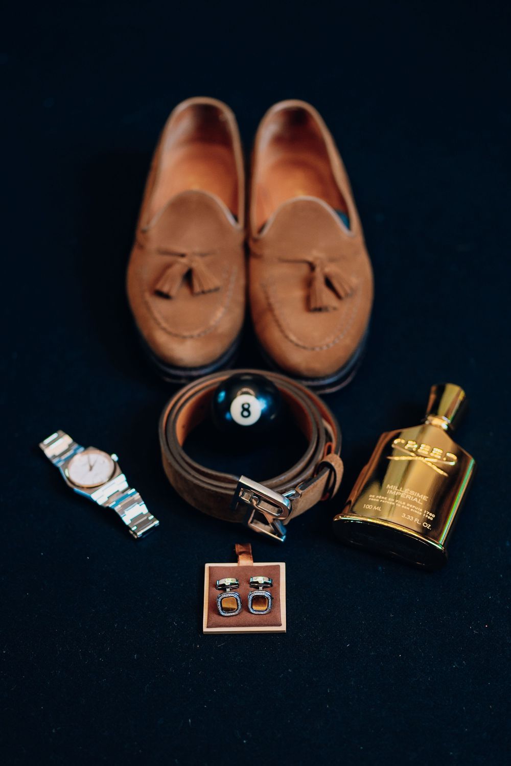 Groom's accessories including brown leather loafers, watch, belt, gold perfume bottle and cufflinks on black background.