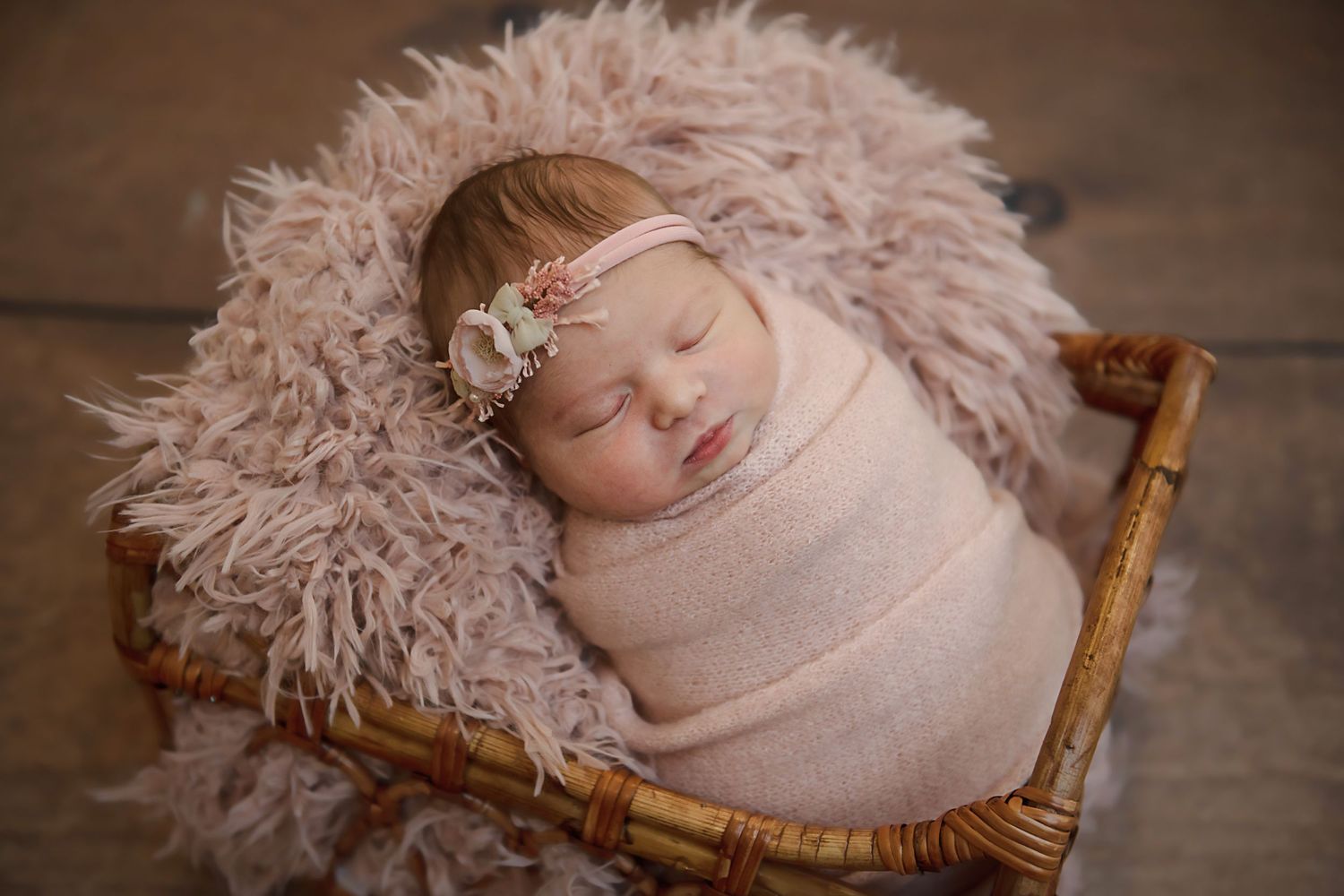 Sleeping newborn baby wrapped in pink swaddle on fluffy blanket in wooden basket.