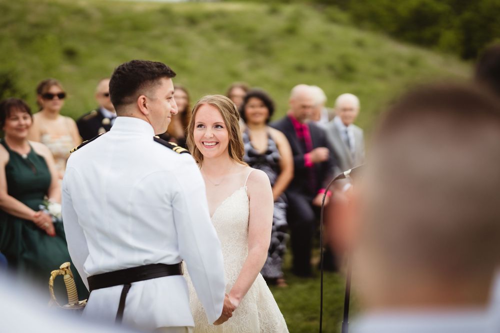 A couple exchanges joyful smiles during their outdoor wedding ceremony with guests looking on in the background.