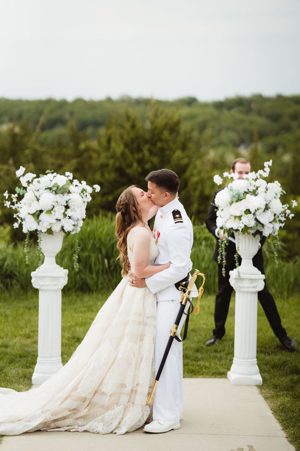 A couple shares their first kiss at an outdoor wedding ceremony between elegant white floral arrangements on pedestals.
