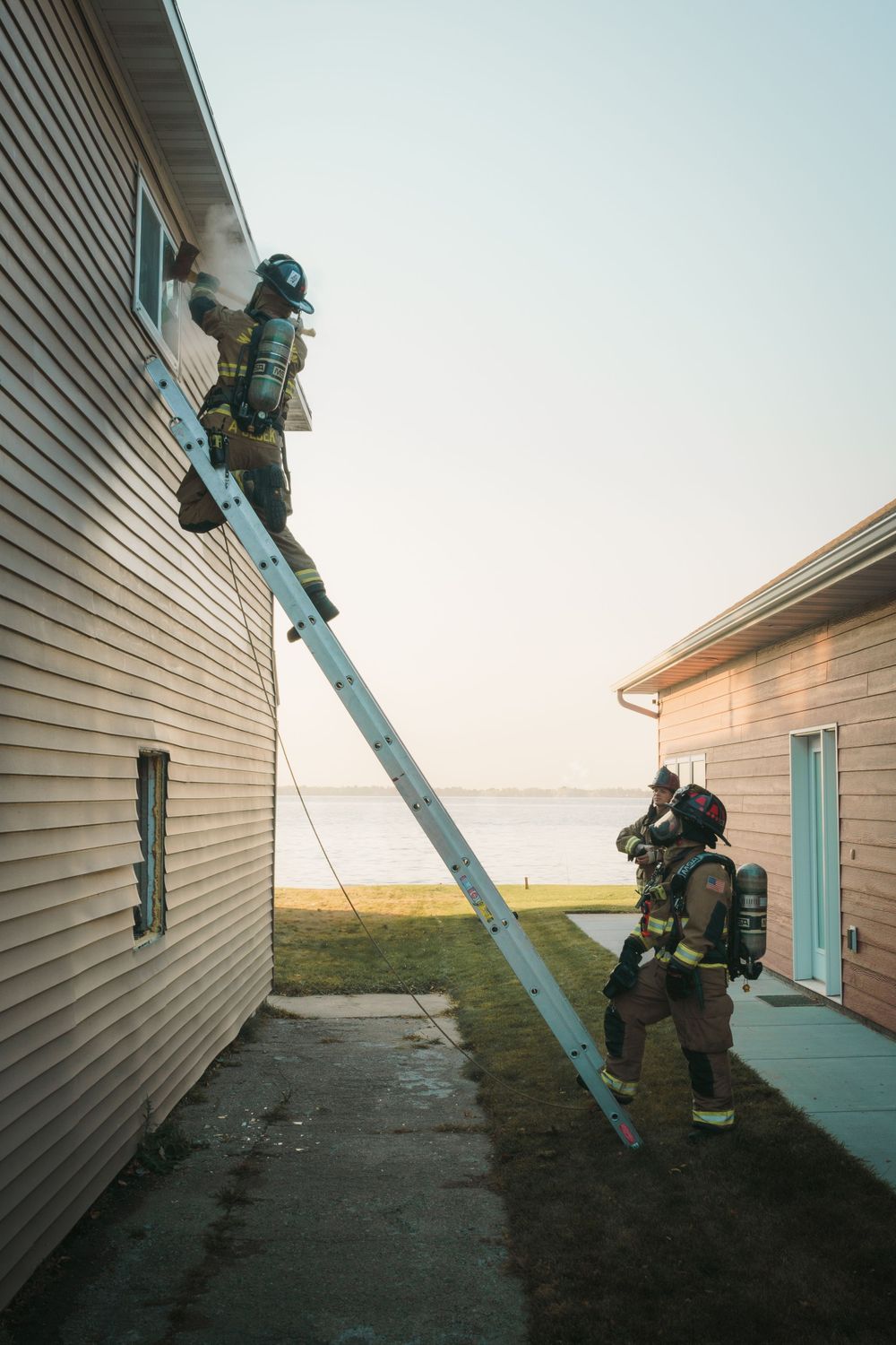 Firefighters in full gear work together using a ladder against a house exterior with water visible in background.