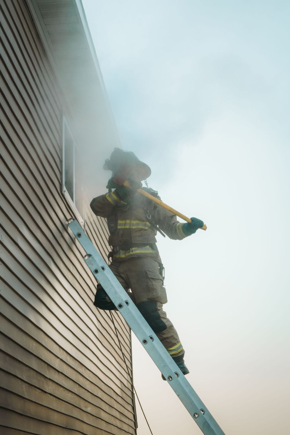 Firefighter climbing a ladder against a house exterior during a rescue operation in smoky conditions.