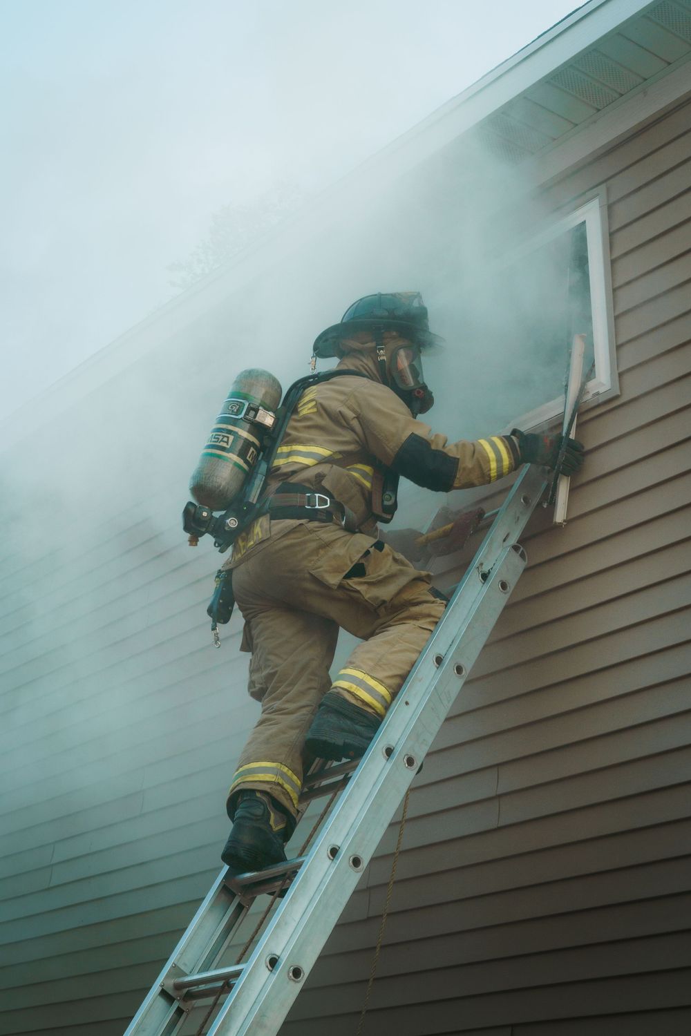 A firefighter in protective gear climbs a ladder through smoke while responding to an emergency at a residential building.