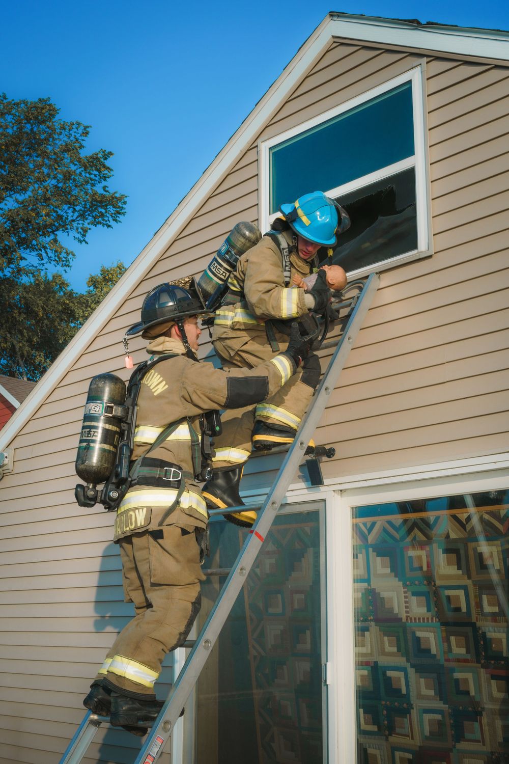 A firefighter in full protective gear climbs a ladder against a beige house facade while carrying an oxygen tank.