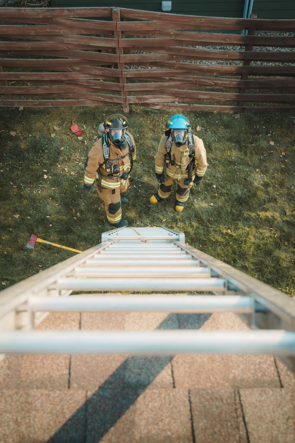 Firefighters in protective gear stand below an extended ladder against a wooden fence during training exercise.