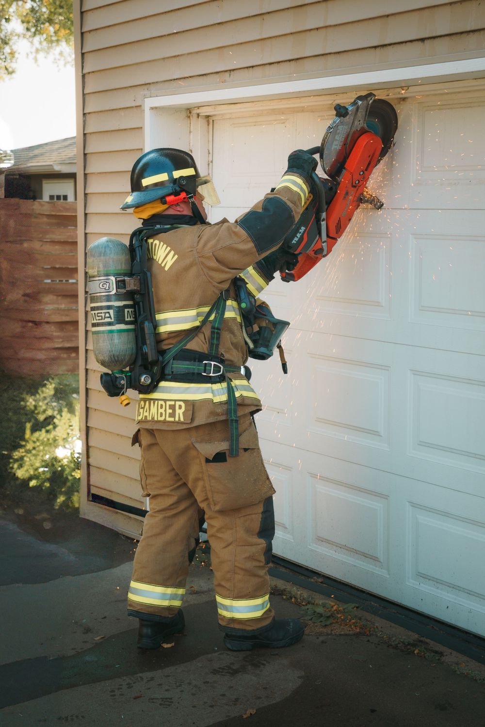 Firefighter in protective gear using a chainsaw on a white garage door during training or maintenance work.