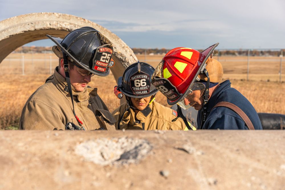 Three firefighters in protective gear and helmets huddle together while working on an incident in a field.