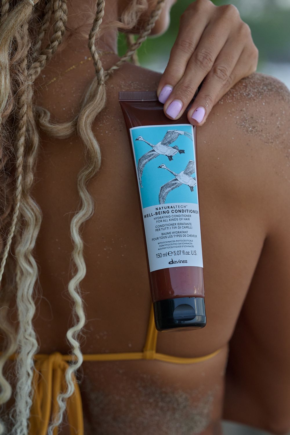 Hands with lavender nails holding Davines hair product with blue label against blonde braided hair.