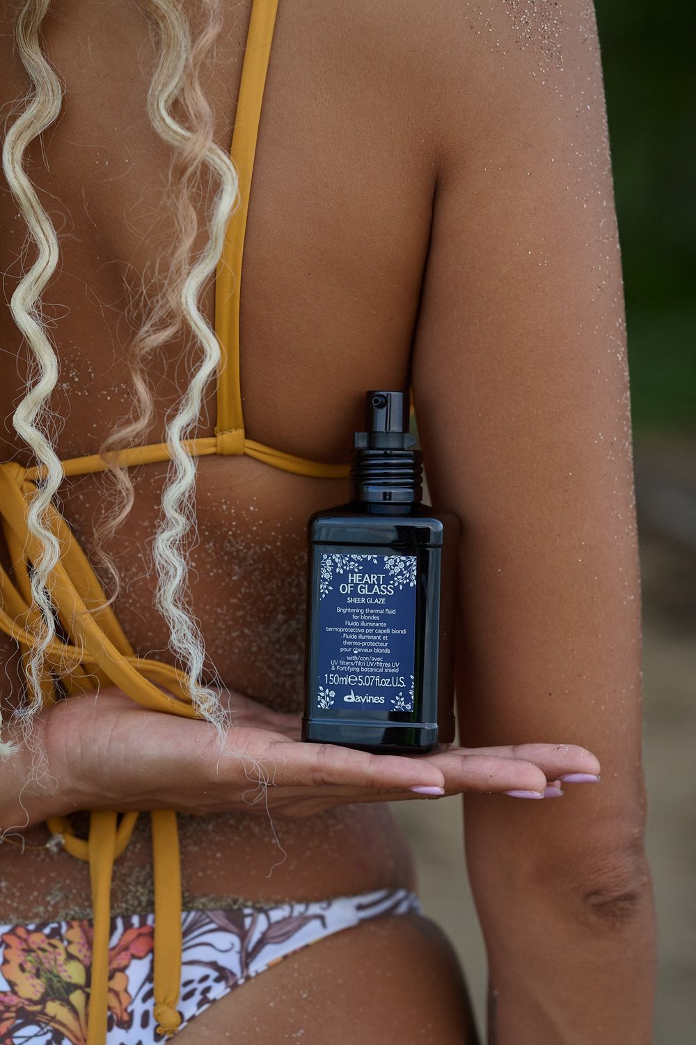 A person in yellow swimwear holds a small dark bottle of tanning oil against tanned skin with glittery textures.