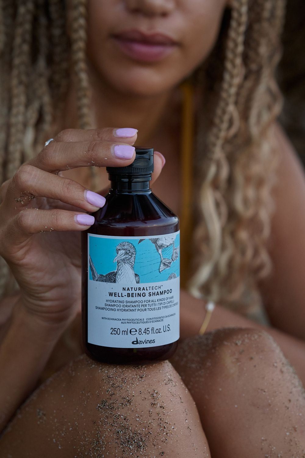 Close-up of hands holding a blue-labeled Davines haircare bottle against a body with blonde braids and glitter.