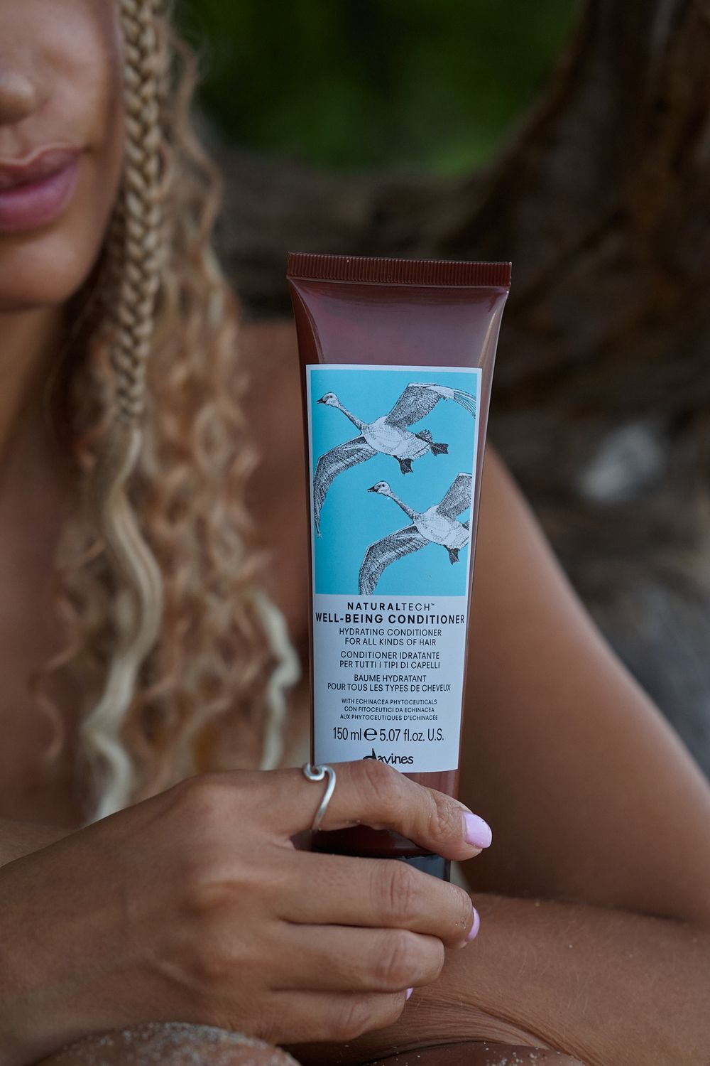A hand holds a blue skincare tube with bird designs against a blurred natural background.