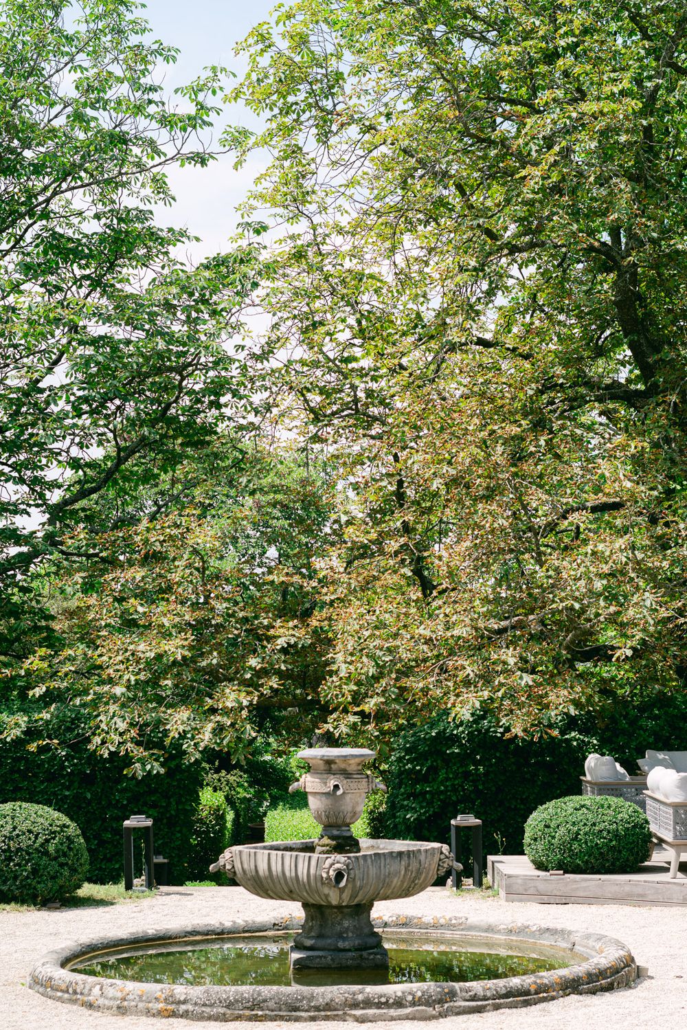 Elegant stone fountain surrounded by lush greenery and manicured hedges in a formal garden setting.