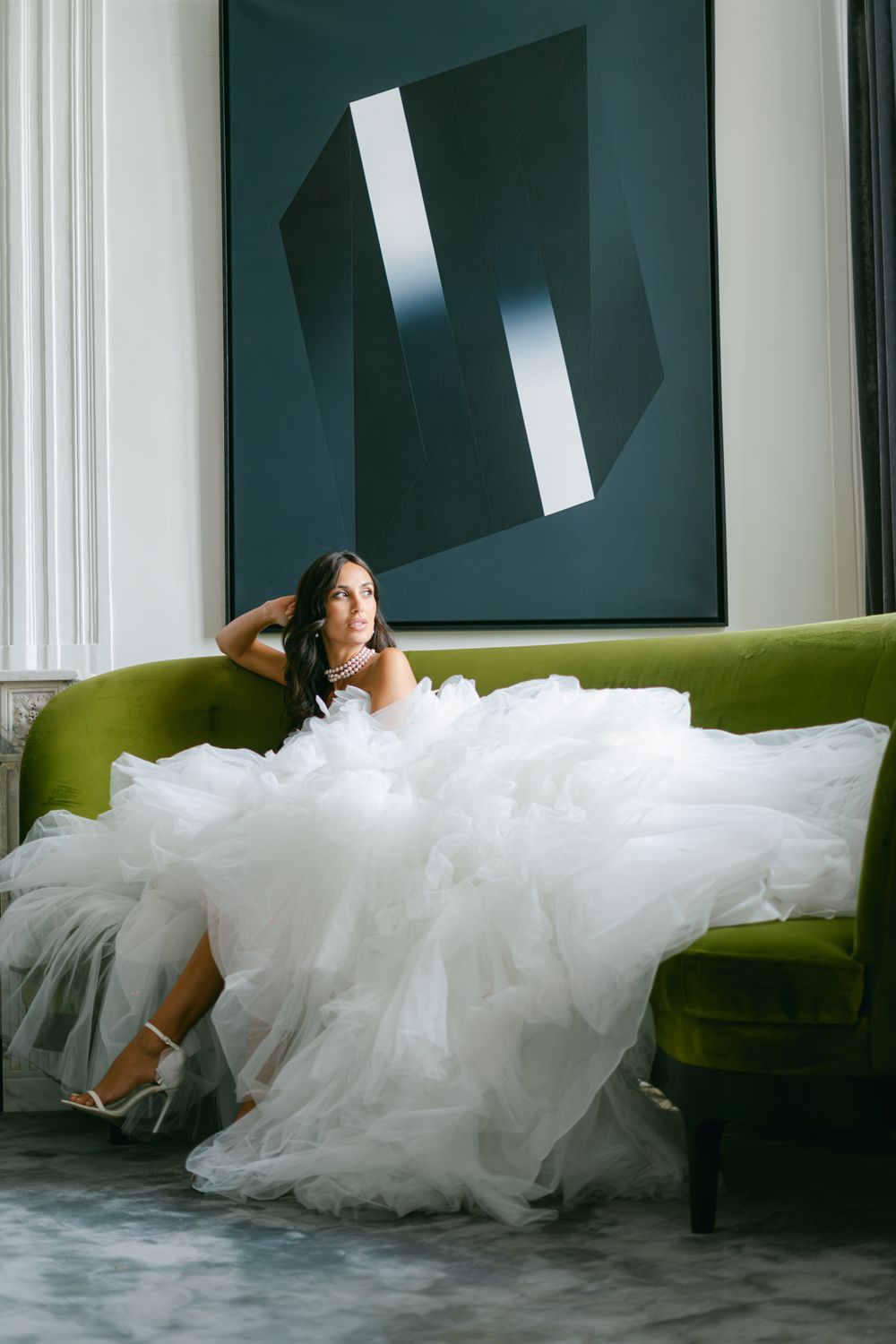 A flowing white wedding dress draped over a green velvet couch beneath modern wall art.
