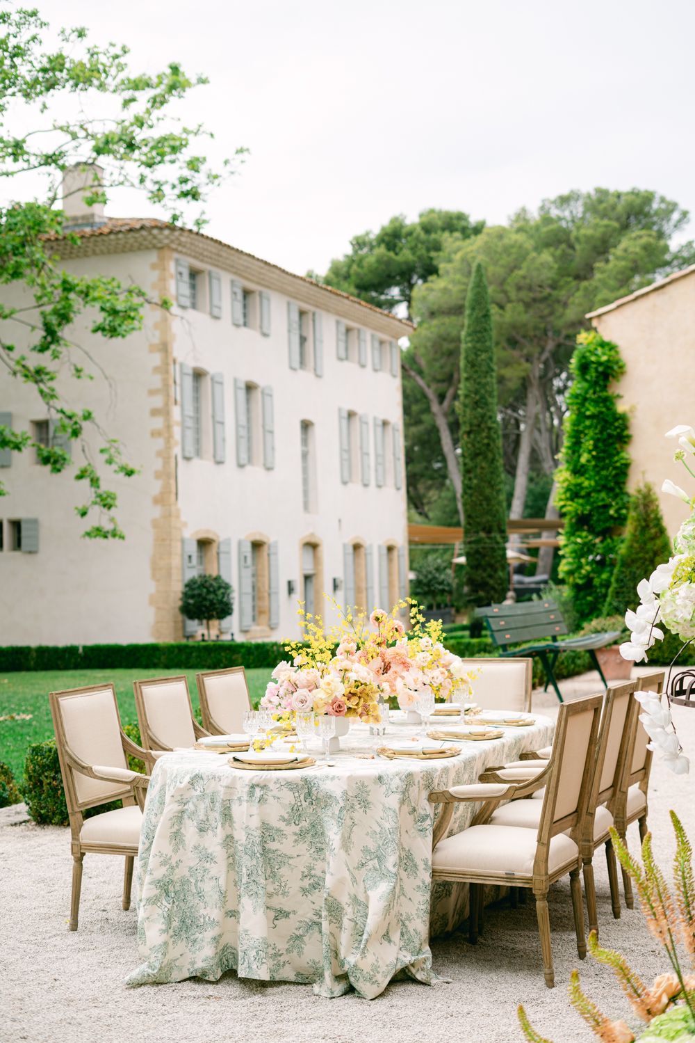 Elegant outdoor wedding table setting with floral tablecloth and cream chairs near a French villa.