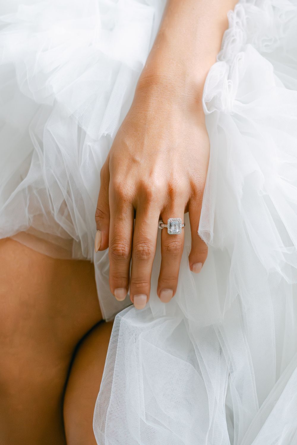 Close-up of diamond engagement ring on hand against white tulle dress fabric.