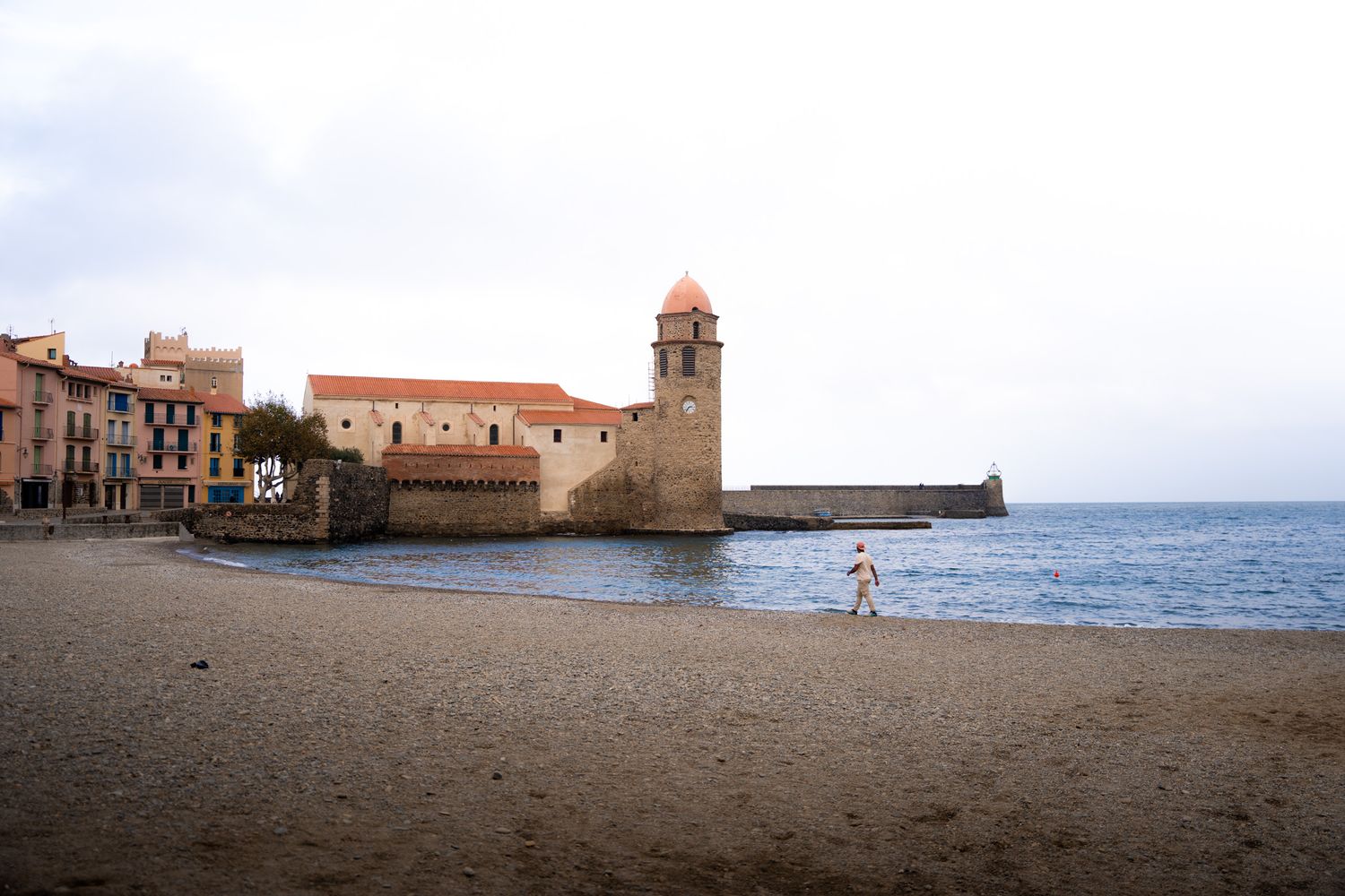 Promeneur sur la plage principale de Collioure avec les clocher en arrière plan et les maisons colorées