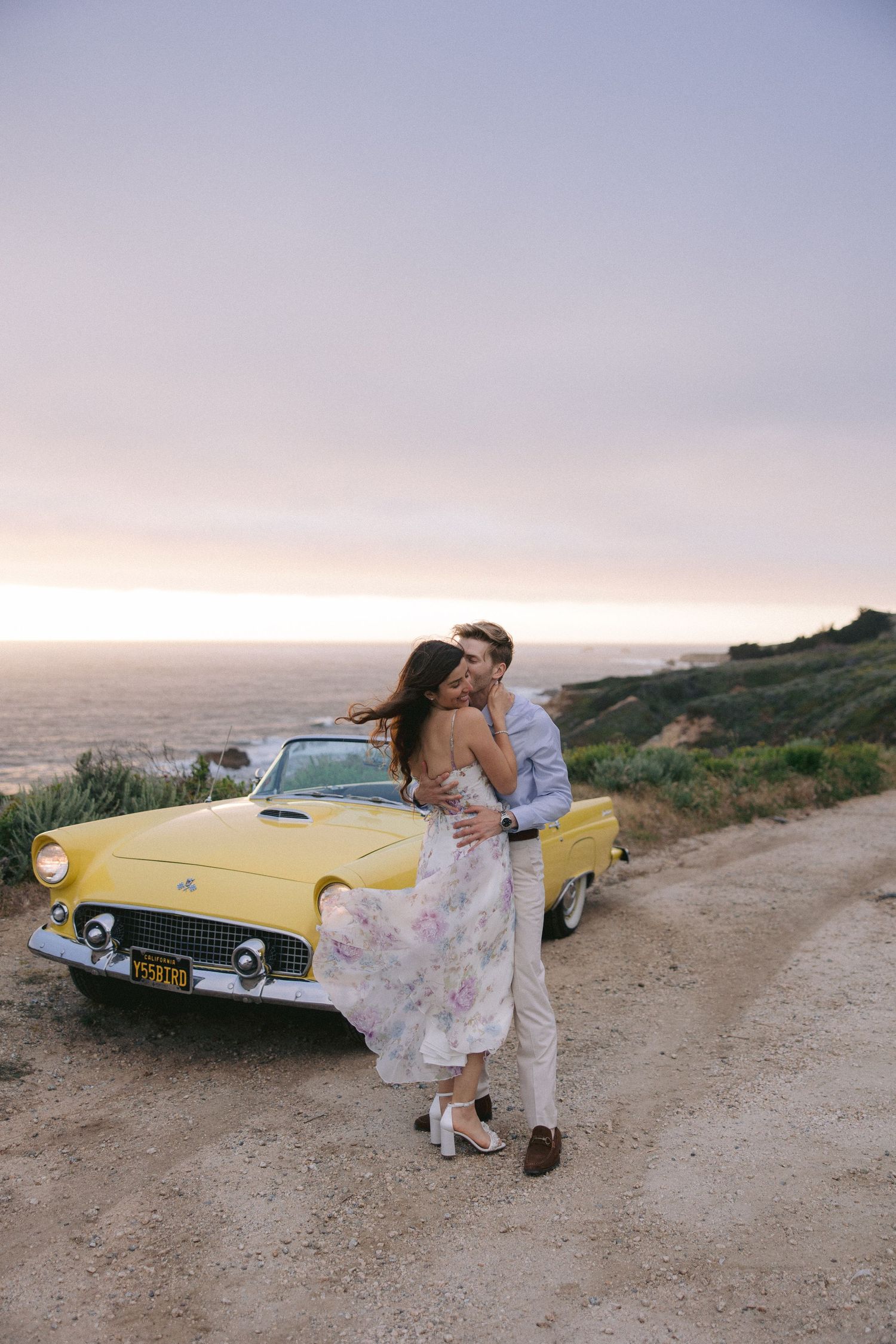 Vintage yellow Thunderbird convertible parked on a scenic coastal road during sunset with couple sharing romantic moment.