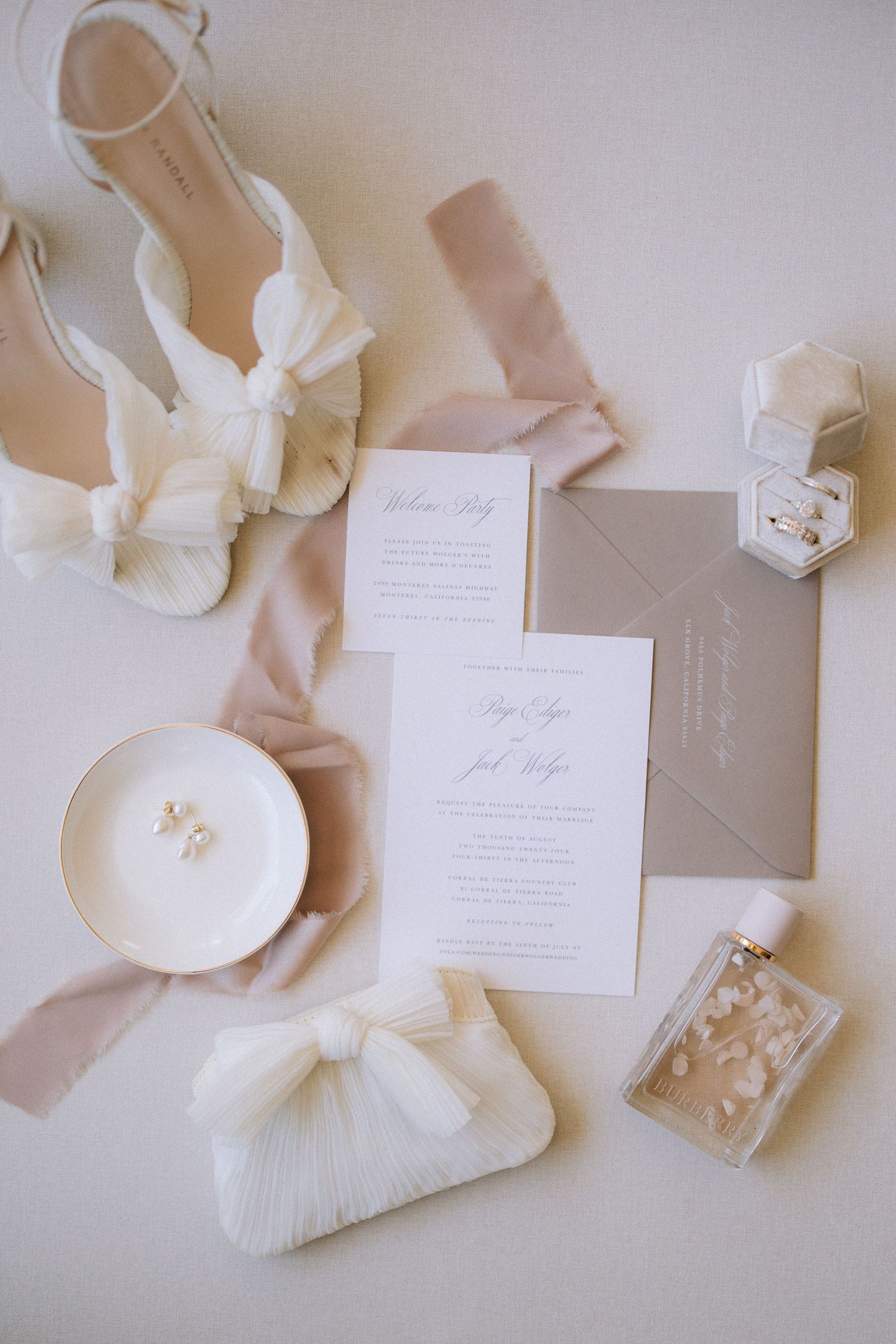 Elegant wedding accessories including white bridal shoes, invitation suite, perfume bottle and clutch arranged on light background.
