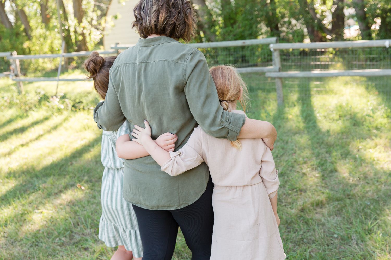 Three people embrace while standing together in backlit garden setting.