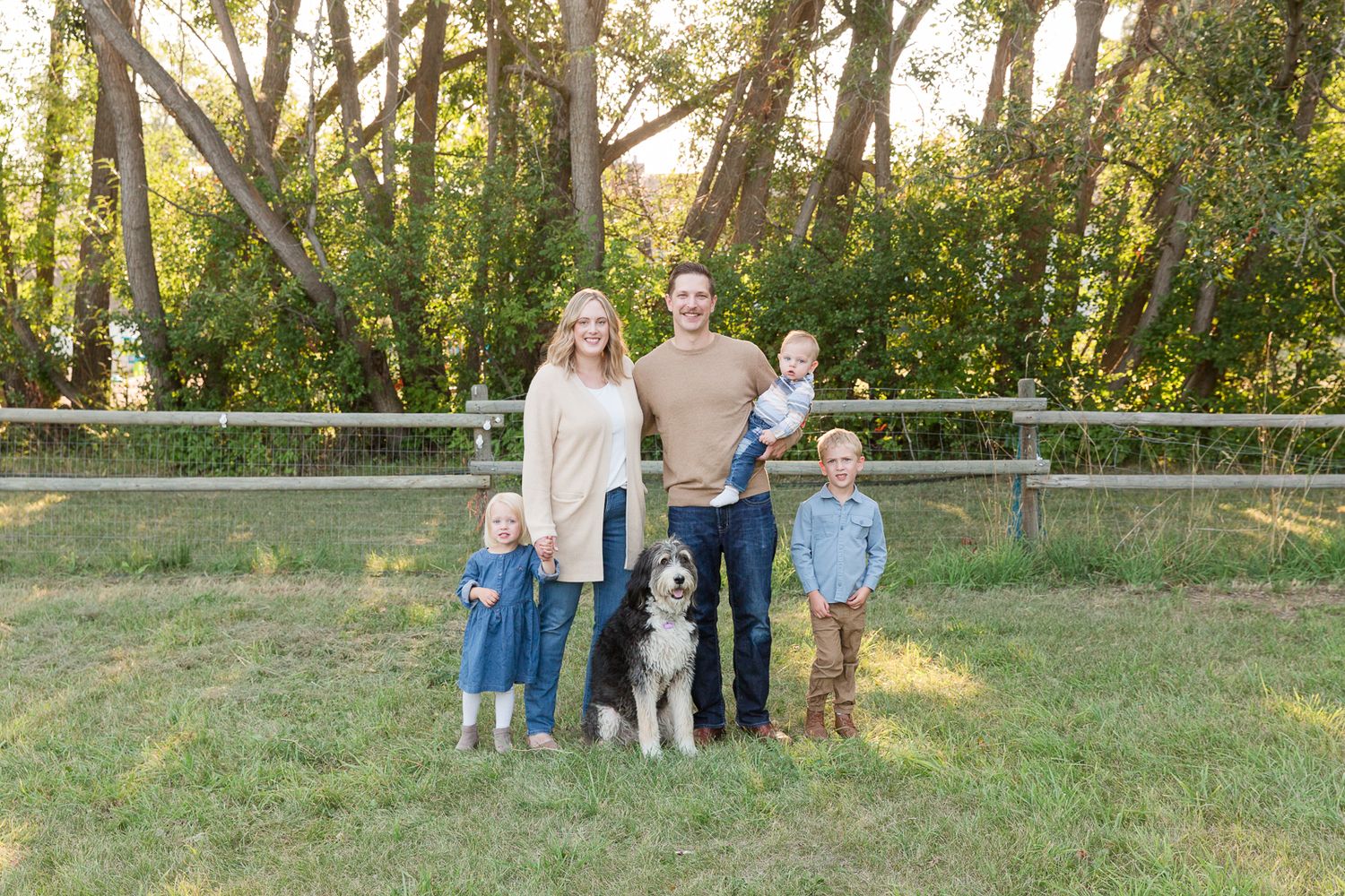 Family poses outdoors with their black and white dog against a rustic fence and trees.