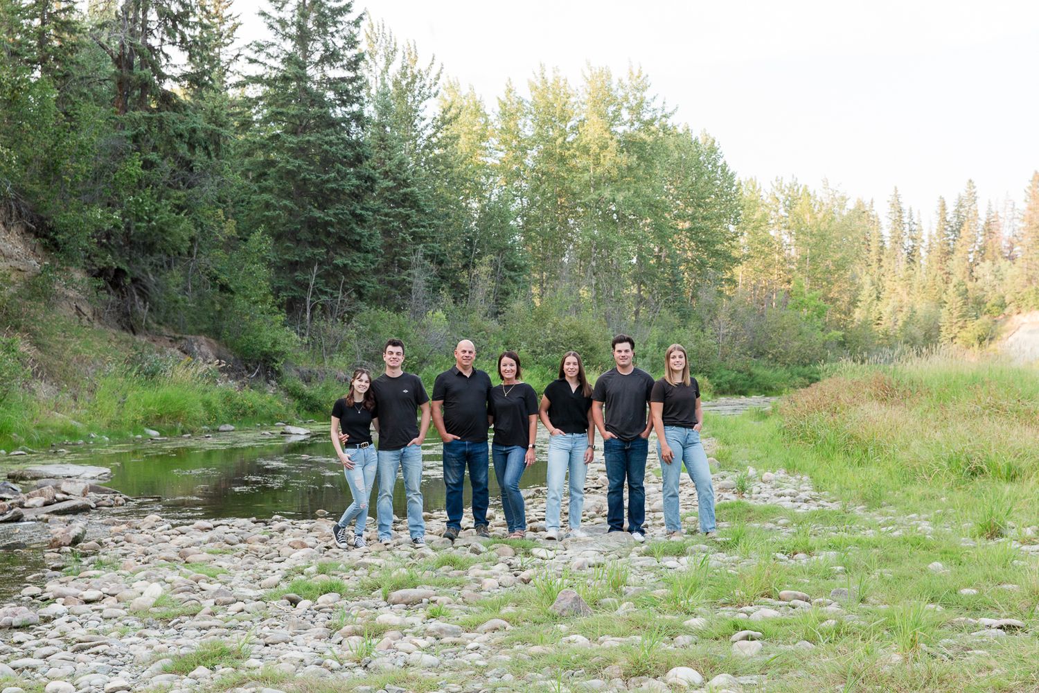 Group stands together by a creek with evergreen trees and sunlit forest in the background.