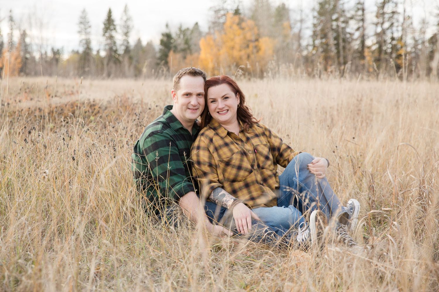 Couple sitting together in tall autumn grass wearing plaid shirts.