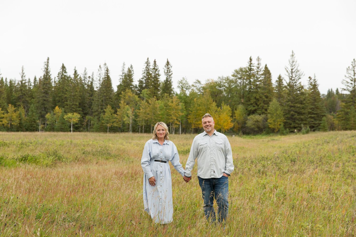 Couple walking through meadow with evergreen forest backdrop.