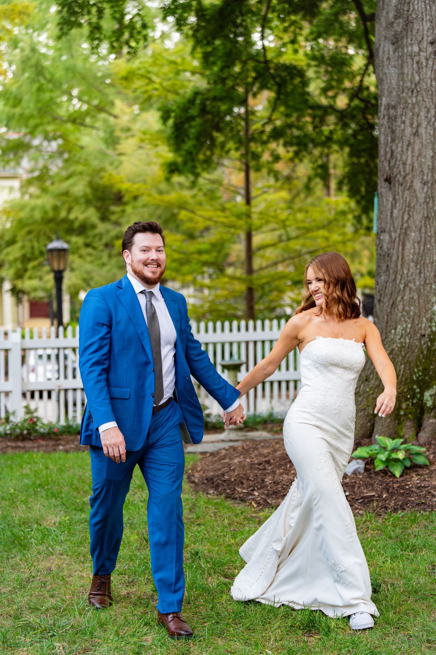 A couple in wedding attire holds hands while walking through a garden with a white picket fence in the background.