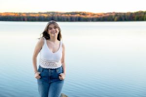 client in white top and jeans poses casually by serene lake during sunset.