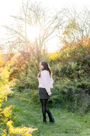 client in white sweater and plaid skirt poses against golden sunset in nature.
