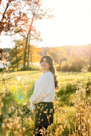 client in cream sweater and plaid skirt stands in golden field during sunset.