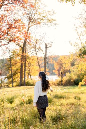 Silhouette in white sweater stands in autumn field with golden light streaming through trees.