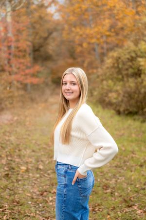 A client in a cozy white sweater poses outdoors against colorful fall foliage.
