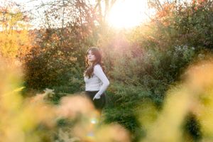 A client in a white sweater stands among autumn foliage during golden hour with sunlight streaming through the trees.
