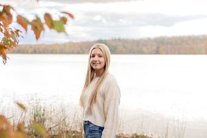 A client in a cream sweater smiles while standing by a lake during autumn.