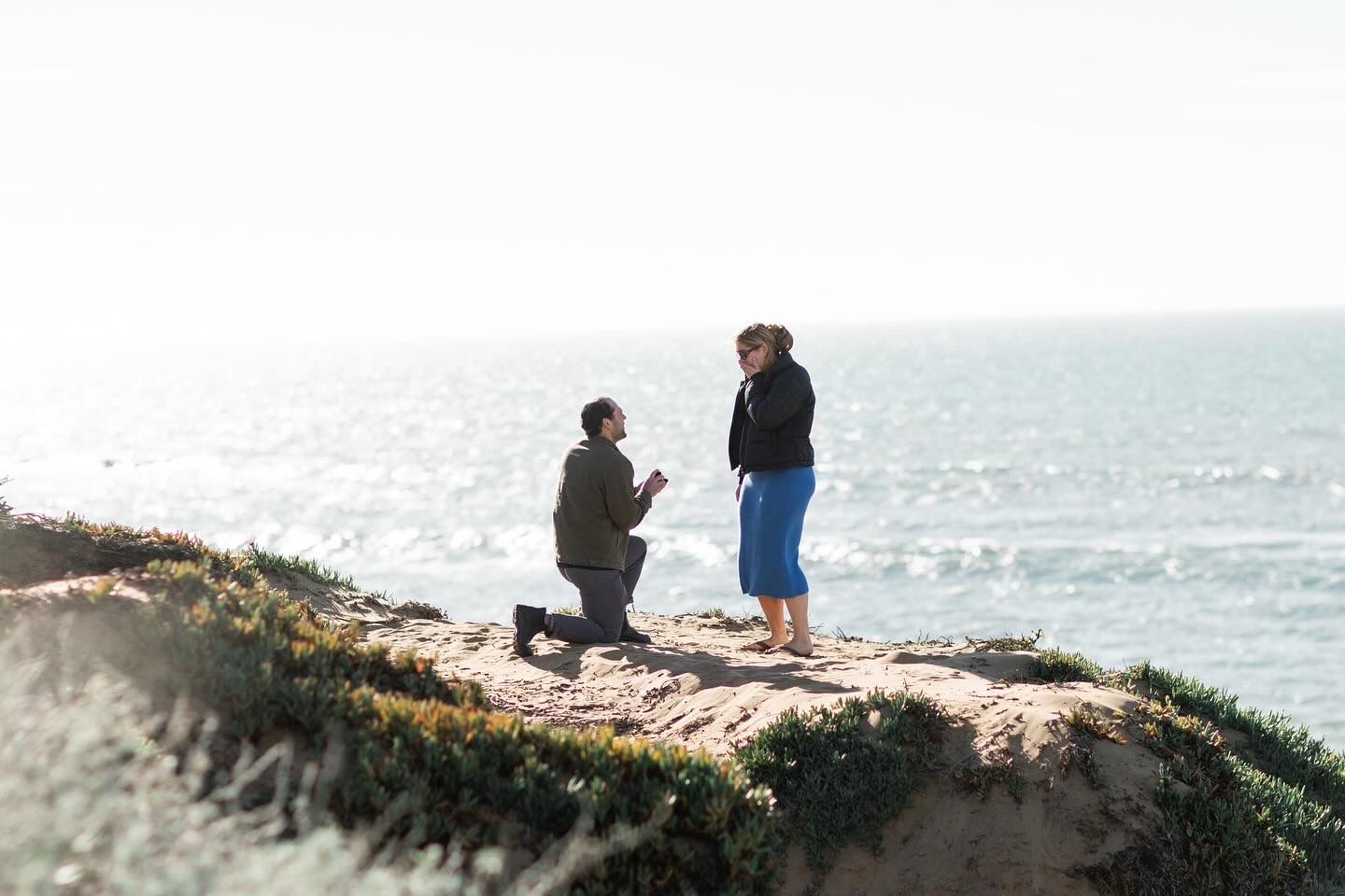 A heartfelt proposal on a scenic coastal cliff overlooking the ocean on a bright sunny day.