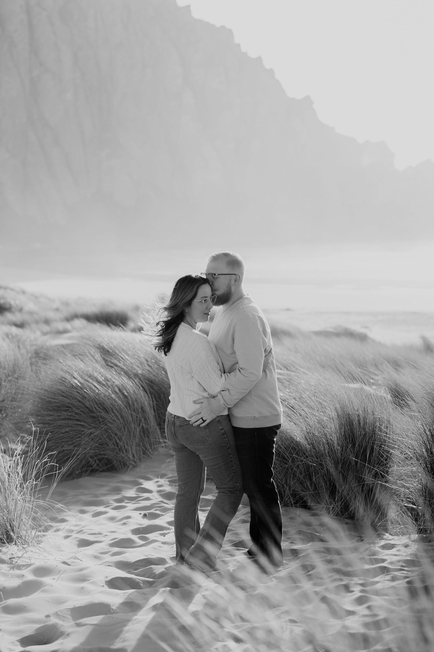 Couple embracing in misty beach dunes with tall grass and moody black and white tones.