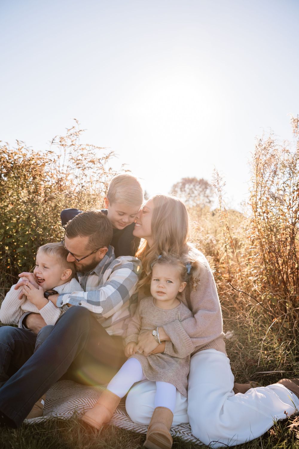 Family of four sitting outdoors in golden sunlight, parents kissing children among tall autumn grasses.