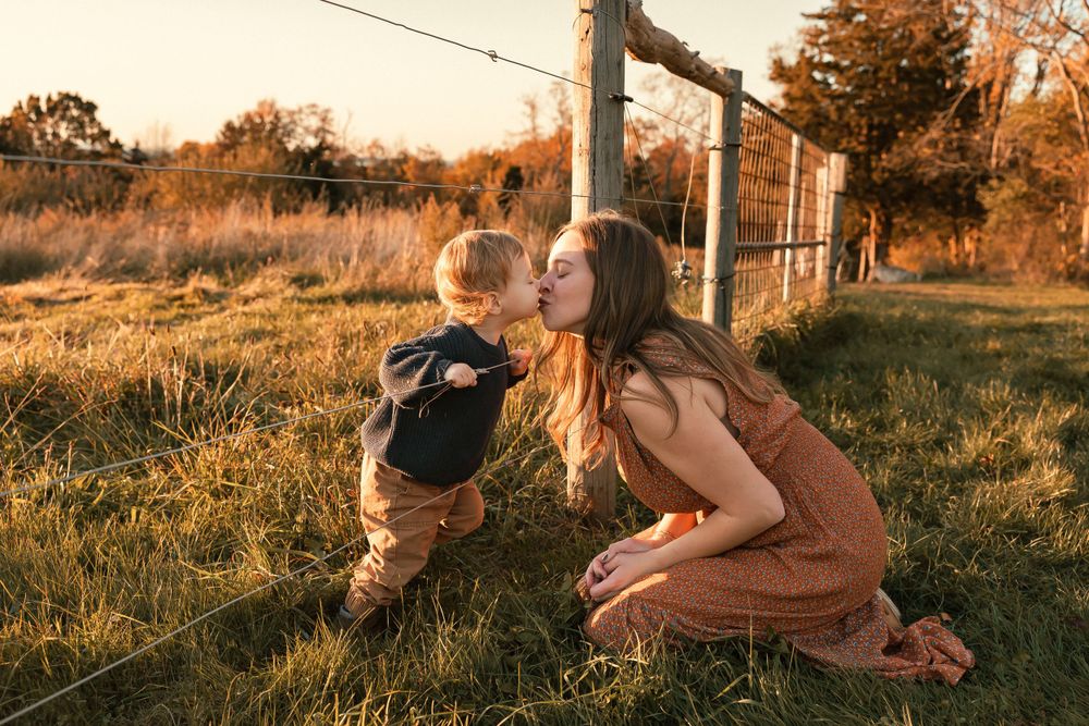 Two people share a tender moment in a sunlit field at golden hour with rustic fence posts in the background.