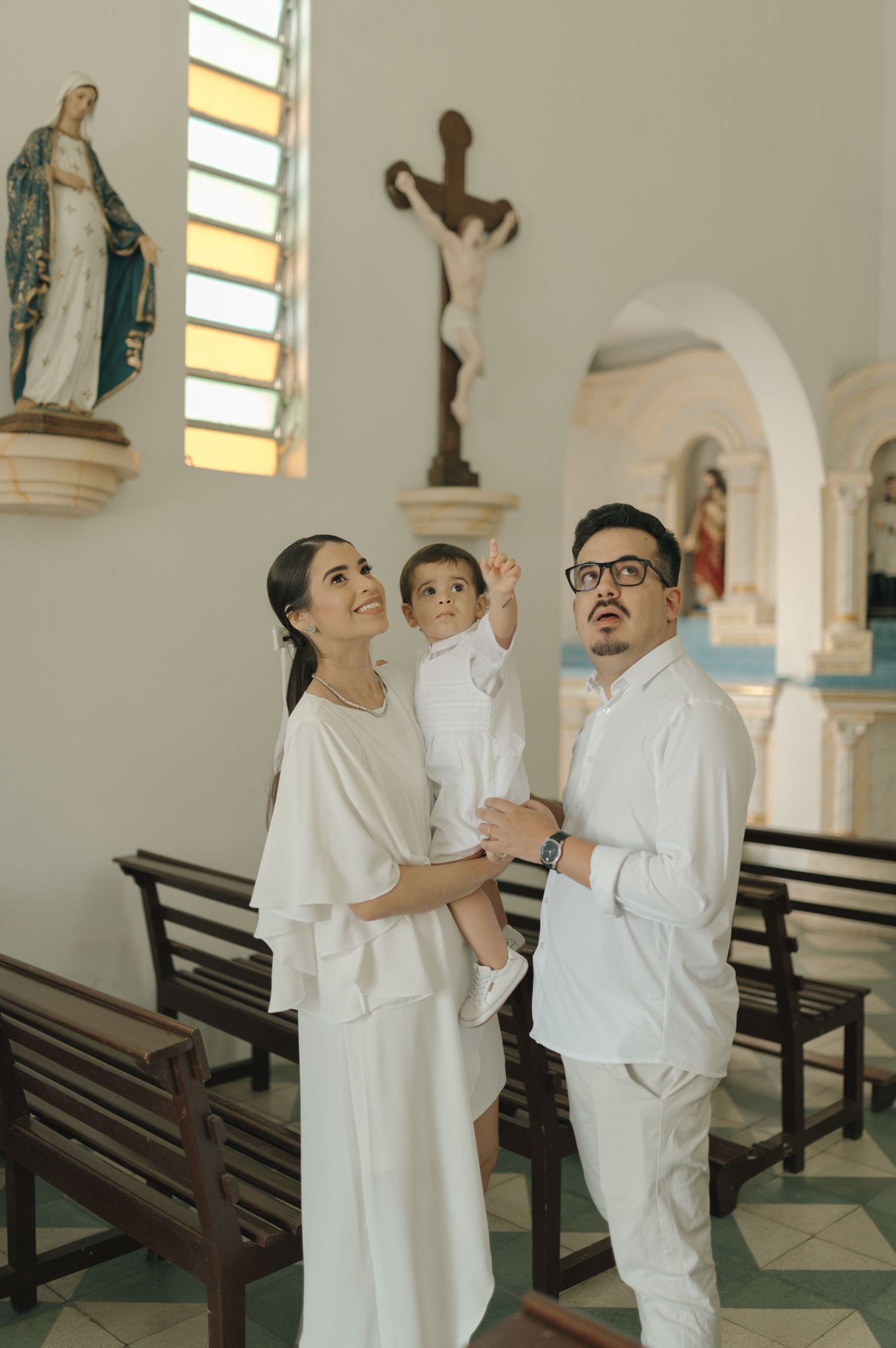 A family in white attire stands together in church, looking up at religious statues and decorations.