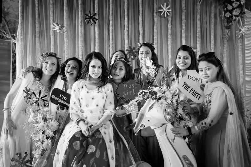A joyful bridal party in pastel dresses poses together holding bouquets against a white backdrop with hanging decorations.