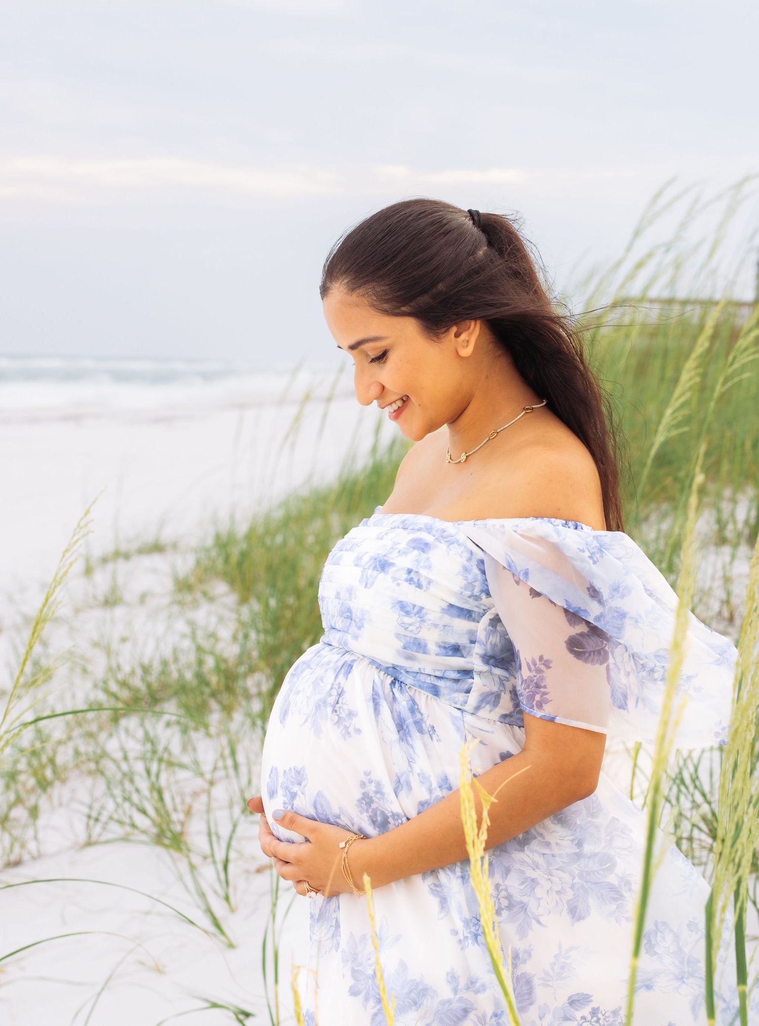 Woman in blue and white dress standing among beach grass near ocean.