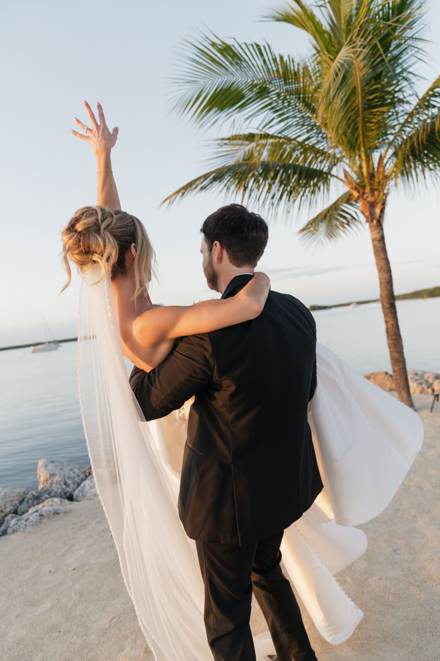 Bride and groom embrace on tropical beach at sunset near palm tree, bride raising arm with flowing veil.